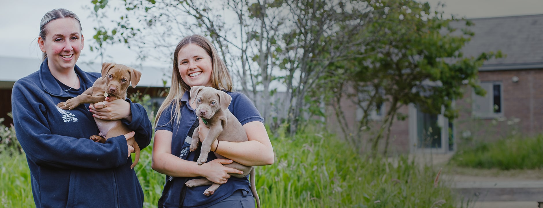 Two female staff members at the Cotswolds Dogs and Cats home holding two puppies outside of the charities building