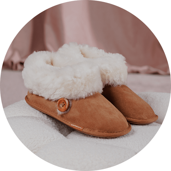 A pair of chestnut Cotswold Wotton Sheepskin Bootsie Slippers on a white cushion