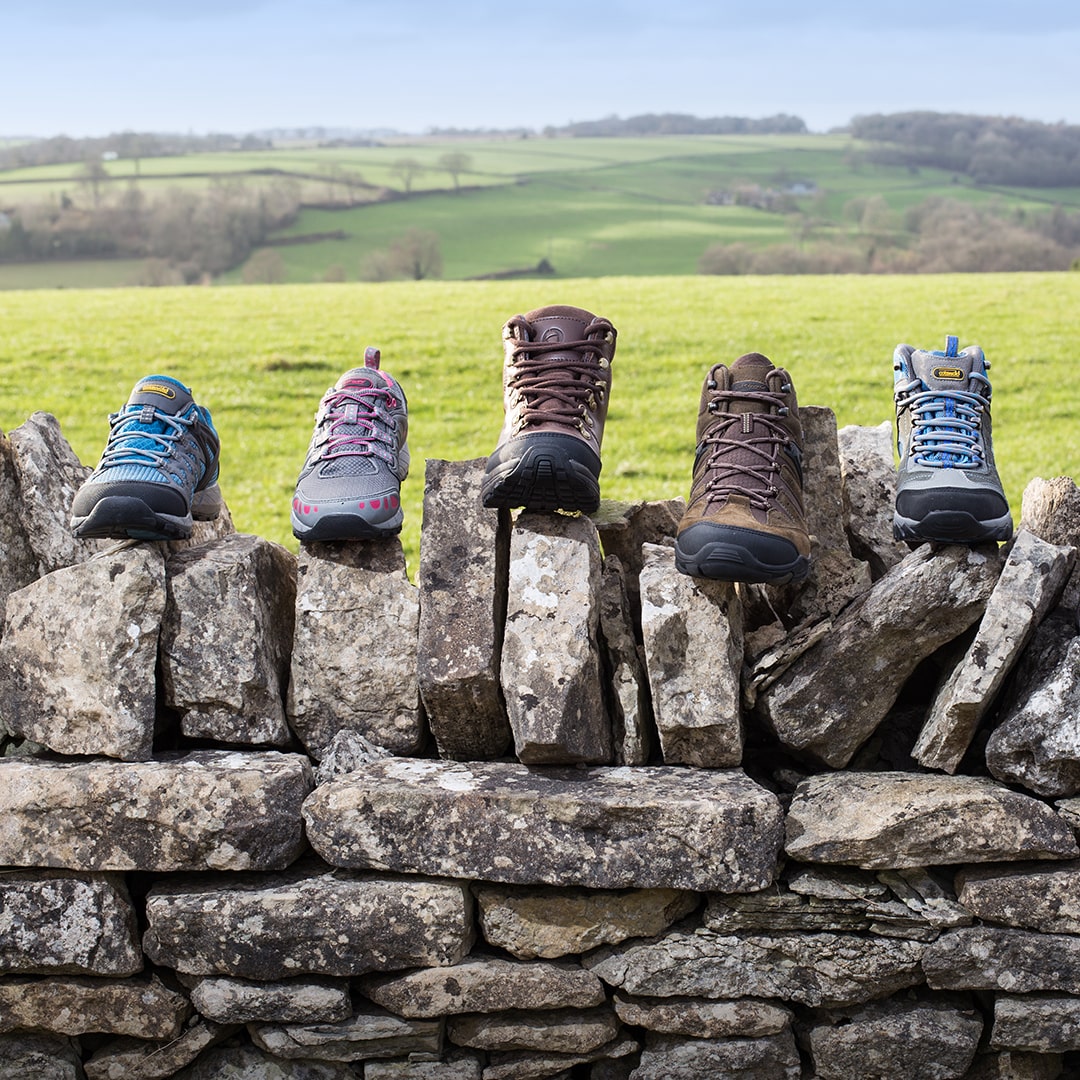 A selection of Cotswold walking shoes and boots on a dry stone wall with rolling hills in the background