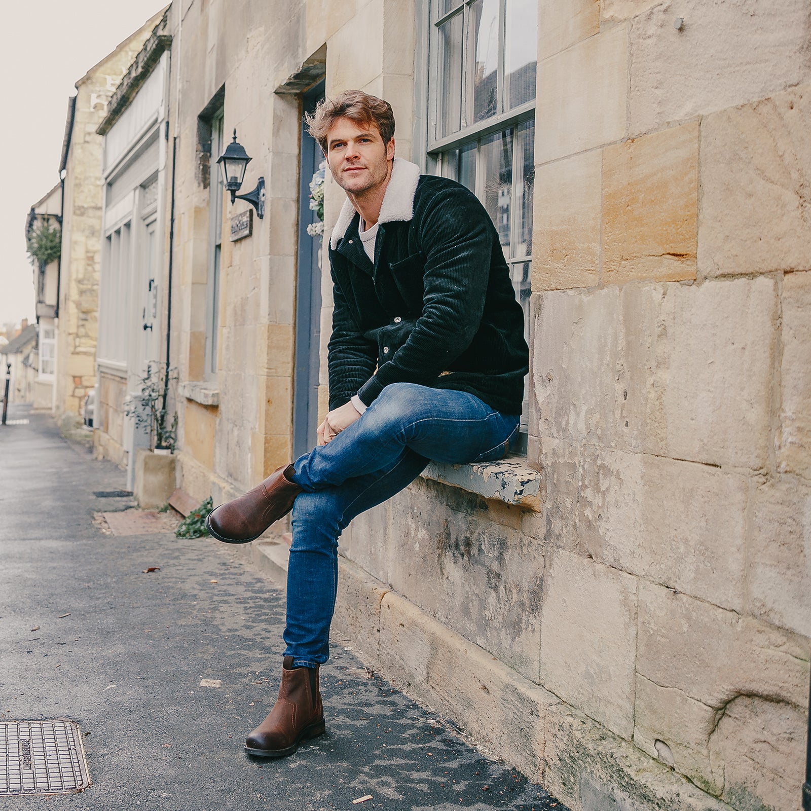Man sat on a windowsill in a Cotswold town, wearing a pair of brown Cotswold Dealer Boots