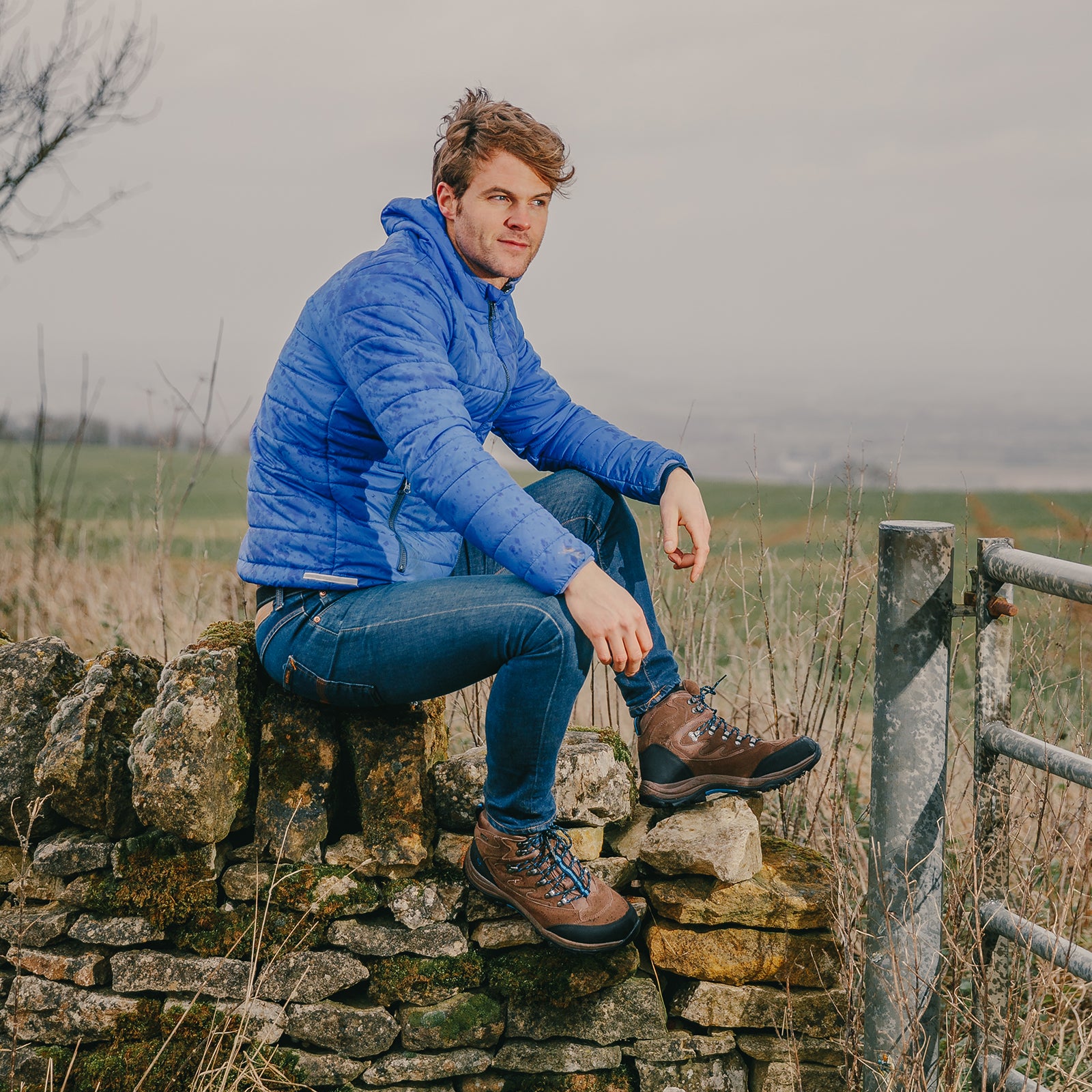 Man sat on a dry stone wall, wearing a blue jacket and a pair of brown Cotswold Oxerton Waterproof Hiker boots