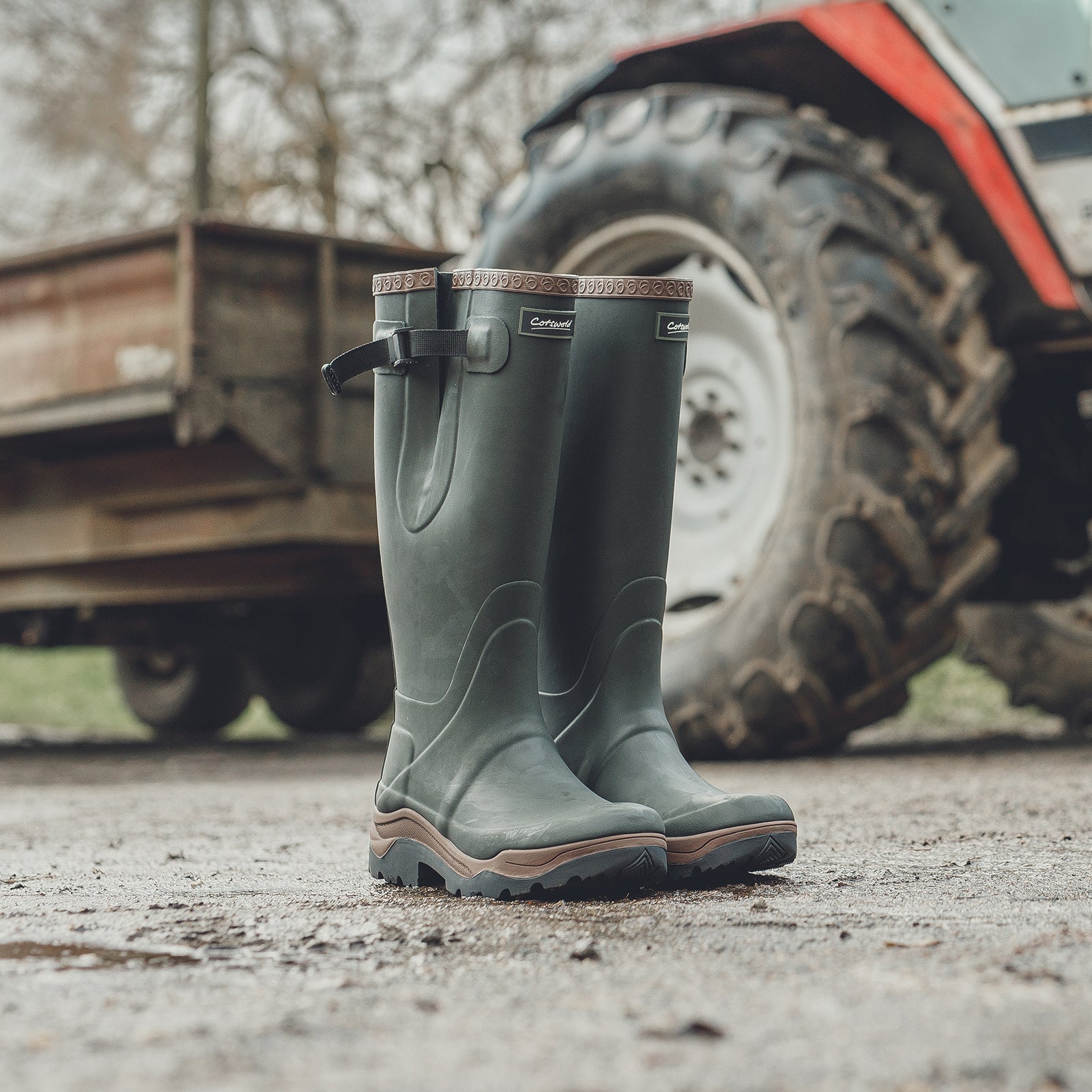 A pair of green Cotswold Compass Wellingtons on muddy ground with a tractor behind