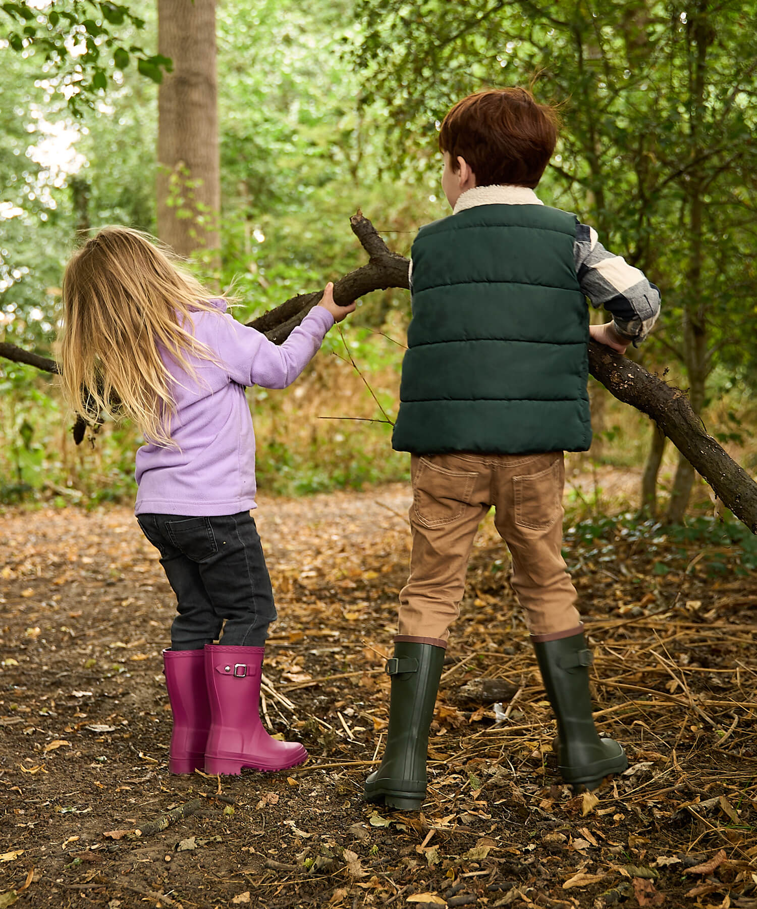 A little boy and girl holding a tree branch with both of them wearing Cotswold wellingtons