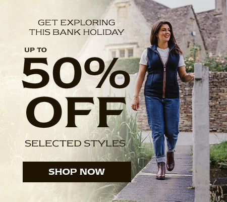 Woman wearing a blue gilet, jeans and pair of Cotswold boots walking along a small bridge with reeds to the side. Text reads 'GET EXPLORING THIS BANK HOLIDAY. UP TO 50% OFF SELECTED STYLES'