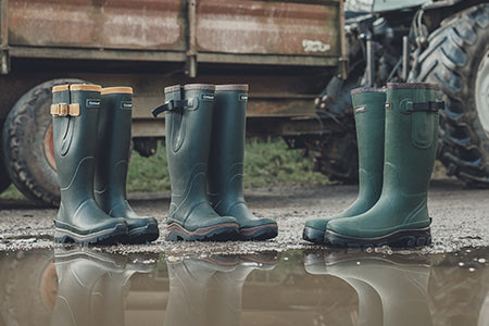 Three pairs of Cotswold wellingtons with reflections in the water and a tractor in the background